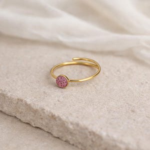 Puede incluir: Un anillo dorado con un centro circular rosa y punteado. El anillo tiene un diseño delicado y abierto con una banda que se envuelve alrededor del dedo. El anillo se encuentra sobre un fondo neutro, resaltando su elegancia.