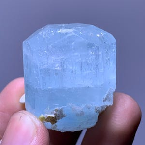 May include: A light blue aquamarine crystal with a hexagonal top. The crystal has a translucent appearance and is held in a hand. The base of the crystal has a rough, textured surface with some brown and yellow coloration.