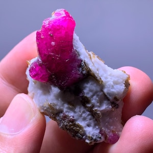 Op de afbeelding: Een ruw, levendig magentakleurig kristal ingebed in een ruwe, witte en grijze mineraalmatrix. Het kristal heeft een doorschijnende, grillige structuur met dieproze tinten, die contrasteert met de getextureerde, ondoorzichtige stenen basis.