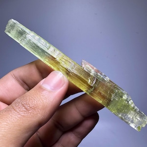 May include: A translucent, elongated crystal with a pale yellow-green hue. The mineral specimen has a rough, textured surface and a slightly fractured appearance. The crystal is held in a hand, showcasing its natural form and color variations. The crystal is a mineral specimen.