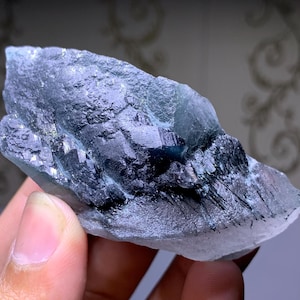 Può includere: Primo piano di un campione minerale grezzo e naturale. La pietra presenta un mix di strutture cristalline blu-verdi e nere. La consistenza ruvida e le diverse sfumature creano un display visivamente interessante. Il minerale è tenuto in una mano.