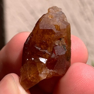 Puede incluir: Un espécimen de cristal en bruto translúcido de color ámbar con una superficie dentada y multifacética. La piedra presenta tonos naranja oscuro y marrón con inclusiones internas visibles y bordes afilados e irregulares.