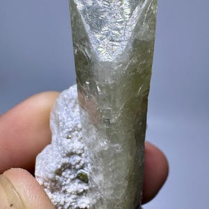 Op de afbeelding: Een doorschijnend, lichtgroen kristal met een rechthoekige vorm, gemonteerd op een witte, getextureerde basis. Het kristal heeft een licht taps toelopende vorm en een reflecterend oppervlak, met natuurlijke minerale formaties. Het geheel suggereert een ruwe, onbewerkte edelsteen.