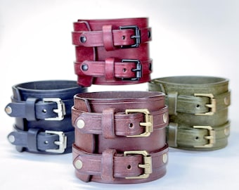 May include: Four wide leather cuff bracelets arranged in a group. One is deep red with black buckles, one is black with silver buckles, one is dark brown with brass buckles, and one is olive green with brass buckles. Each features two straps.