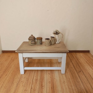 Könnte beinhalten: Ein kleiner Holztisch mit weißem Untergestell und hellbrauner Tischplatte. Darauf stehen kleine Keramikbehälter und ein weißer Krug mit getrockneten Blumen. Die Szene hat eine rustikale, Vintage-Ästhetik.