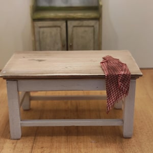 Puede incluir: Una pequeña mesa de madera con tapa de color claro y patas blancas. Un paño a cuadros rojos y blancos está drapeado sobre el borde. Un armario verde con tazas blancas cuelga en el fondo, lo que sugiere una cocina o un comedor.