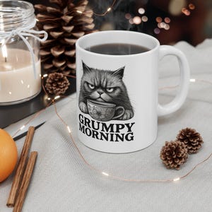 May include: White ceramic mug with a grumpy cat design holding a teacup. The mug has the words "GRUMPY MORNING" printed below the cat. The mug is filled with a dark liquid.