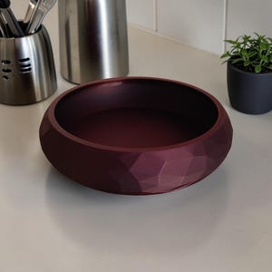 May include: A burgundy-colored, faceted bowl with a geometric design. The bowl is round and sits on a white surface. The bowl is empty and appears to be made of ceramic or a similar material. Utensil holders and a small potted plant are in the background.