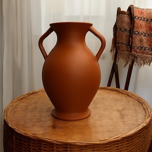 May include: A terracotta-colored vase with two curved handles. The vase has a rounded body and a wide base, sitting on a round, woven table. The vase has a textured surface with horizontal lines. The background includes a curtain and a wooden rack.