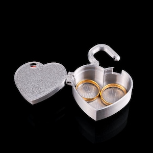 May include: A silver heart-shaped ring box, open to reveal two gold wedding bands. The box has a textured surface and a clasp. The background is black.