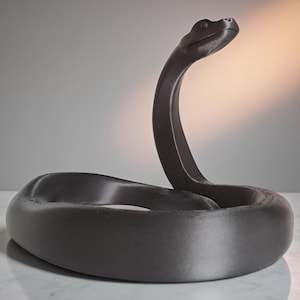 Pode incluir: Uma escultura cinza escuro de uma cobra em posição enrolada. A cabeça da cobra está levantada e o corpo forma uma forma circular. A escultura tem um acabamento liso e fosco sobre um fundo neutro.