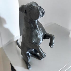 May include: A dark gray sculpture of a dog in a playful pose, standing on its hind legs. The dog's features are detailed, with a focused expression. The sculpture is likely made of a solid material, and the overall aesthetic is modern and minimalist.