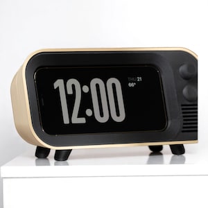 Può includere: Un orologio digitale in stile retrò con uno schermo nero che mostra "12:00" in bianco. L'orologio ha una cornice color oro e dettagli neri, inclusi i piedini e le manopole di controllo. Vengono visualizzati anche la data e la temperatura.