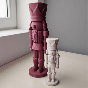 Peut inclure: Deux figurines décoratives de casse-noisettes. L'une est de couleur bordeaux plus grande, et l'autre est de couleur blanc cassé plus petite. Les deux ont un design de soldat classique avec un grand chapeau et reposent sur une base ronde. Les figurines sont posées sur une surface claire.