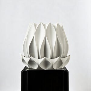 Könnte beinhalten: Eine weiße, abstrakte Keramikskulptur, die einer stilisierten Blume oder Knospe ähnelt. Das Stück weist eine Reihe von gebogenen, blütenblattartigen Formen auf, die sich von einer Basis erheben. Die Skulptur wird auf einem schwarzen, rechteckigen Sockel vor weißem Hintergrund präsentiert.