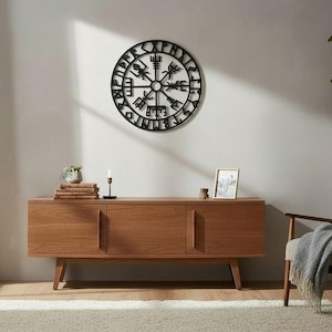 Può includere: Un'opera d'arte murale nera e circolare con simboli intricati e stilizzati è appesa sopra un mobile in legno. Il mobile ha tre ante ed è posto contro un muro neutro. Una sedia con una coperta e una pianta sono sullo sfondo.