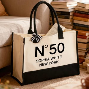 Può includere: Borsa tote color crema con dettagli e manici neri. Il testo "N°50 SOPHIA WHITE NEW YORK" è stampato in nero. Un fiocco a righe bianche e nere adorna un manico.