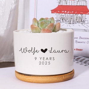 May include: A white ceramic planter with a succulent plant, resting on a small wooden coaster. The planter has the names "Wolfe" and "Laura" with a heart symbol, "9 YEARS 2025" in black script. A second planter is visible.