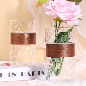 May include: Two clear glass vases with a textured design and a wooden band. One vase holds pink roses. The wooden bands are engraved with names. The vases are on a white surface with a book that says "BLE IN PARIS".