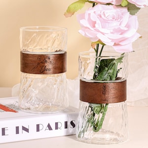 May include: Two clear glass vases with a textured design and wooden bands. One vase has the name "Elisa" engraved, the other has "Paisley". The vases are holding pink flowers. The vases are sitting on a white surface.