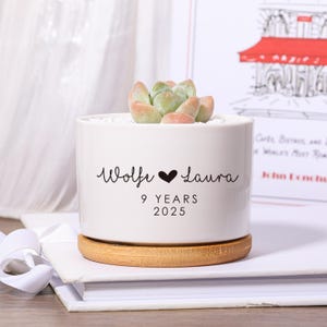 Peut inclure: Un pot de plante en céramique blanche avec une plante succulente, posé sur un dessous de plat en bambou. Le pot porte les noms "Wolfe" et "Laura", un symbole de cœur et le texte "9 YEARS 2025".