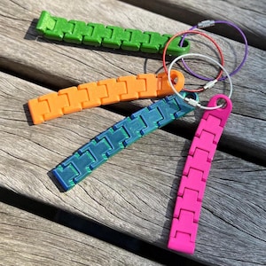 Peut inclure: Plusieurs jouets fidget colorés, verts, oranges, bleus et roses, chacun avec un design segmenté. Ils sont attachés à des porte-clés avec du fil argenté. Le fond est une surface en bois vieilli.