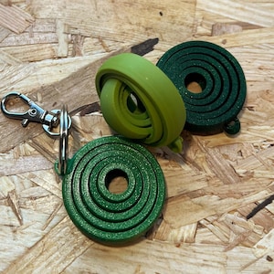 Op de afbeelding: Drie groene, spiraalvormige sleutelhangers en een lichtgroene, opgerolde sleutelhanger. Eén sleutelhanger is bevestigd aan een zilverkleurige sluiting. De sleutelhangers zijn gemaakt van een textuurrijk materiaal en liggen op een houten oppervlak.