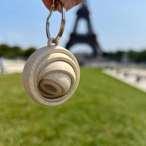 May include: A beige, circular keychain with a spiral design, held in front of the Eiffel Tower. The keychain is made of a speckled material and attached to a silver keyring. The background features green grass and a clear blue sky.