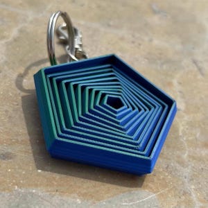 Puede incluir: Un llavero geométrico azul y verde con un llavero plateado. El llavero tiene un diseño en capas, con cada capa disminuyendo de tamaño hacia el centro. El llavero mide aproximadamente 5 cm de ancho.