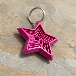 Pode incluir: Um chaveiro rosa brilhante em forma de estrela com um porta-chaves prateado. O design da estrela apresenta múltiplas formas de estrela aninhadas, criando um efeito em camadas. O chaveiro é feito de um material sólido e assenta numa superfície texturizada.