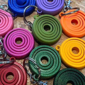 May include: A collection of colorful, circular keychains in shades of pink, purple, blue, green, orange, yellow, and red. Each keychain has a series of concentric rings and a silver keyring. The keychains are made of a solid material.