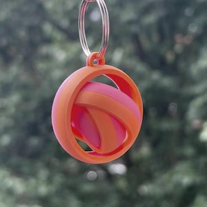 May include: A 3D-printed keyring with a spherical design. It features interwoven orange and pink bands within a circular orange frame. A silver key ring is attached. The background shows blurred green foliage.