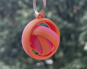 May include: A 3D-printed keyring with a spherical design. It features interwoven orange and pink bands within a circular orange frame. A silver key ring is attached. The background shows blurred green foliage.