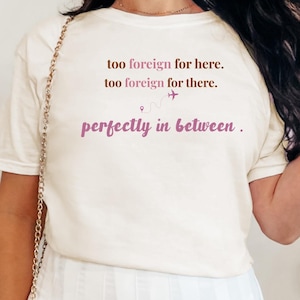 May include: Cream-colored t-shirt with the text "too foreign for here. too foreign for there. perfectly in between." in a mix of brown and purple fonts. The shirt features a small airplane graphic. The shirt is a casual wear item.