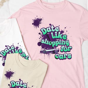 Op de afbeelding: Drie t-shirts in wit, roze en beige, elk met de tekst "Date Like shopping for cars" in een gestileerd lettertype en een autografiek. Paarse verfspatten accentueren het ontwerp.
