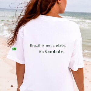 May include: A white short-sleeved t-shirt featuring the black printed text 'Brazil is not a place. It's Saudade.' on the back. A small, rectangular Brazilian flag icon is printed on the left sleeve of the garment, which has a crew neckline.