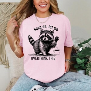 A woman wearing a pink t-shirt with a black graphic of a raccoon waving next to a coffee cup and the text "Hang on, let me OVERTHINK THIS."