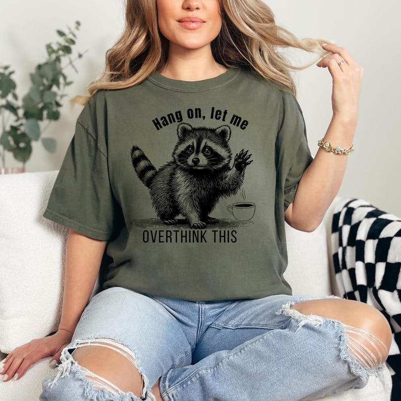 A woman wearing an olive green t-shirt with a black graphic of a raccoon waving next to a coffee cup and the text "Hang on, let me OVERTHINK THIS."