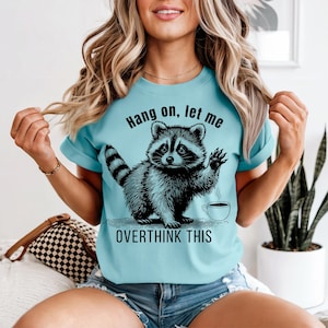 A woman wearing a light blue t-shirt with a black graphic of a raccoon waving next to a coffee cup and the text "Hang on, let me OVERTHINK THIS."