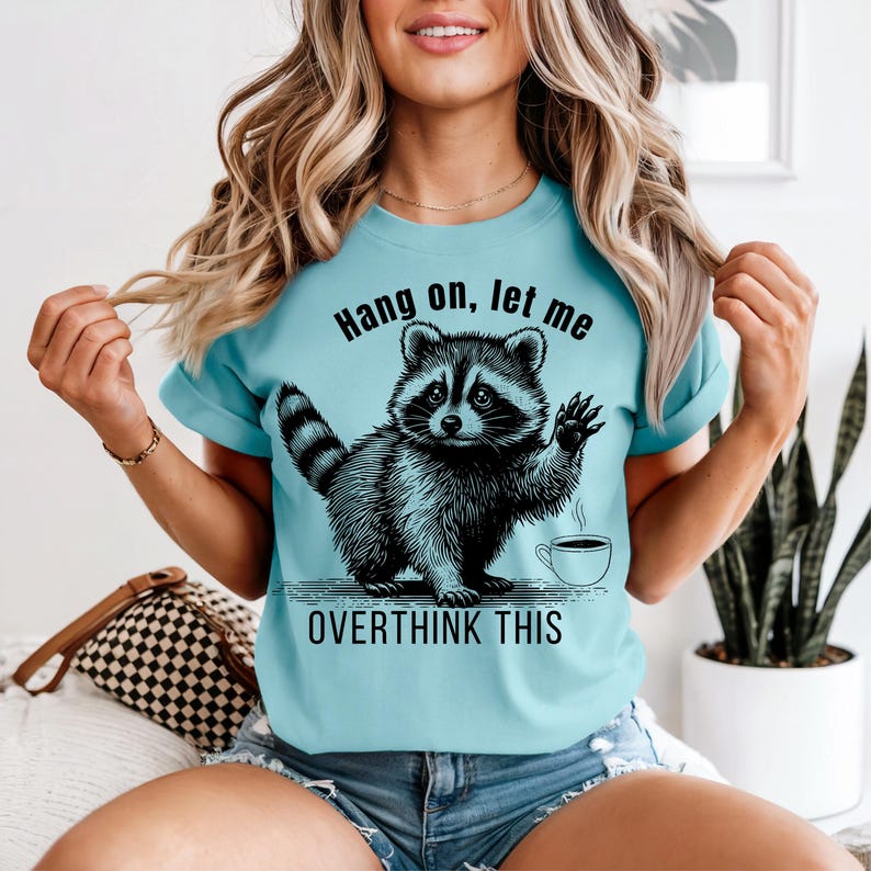 A woman wearing a light blue t-shirt with a black graphic of a raccoon waving next to a coffee cup and the text "Hang on, let me OVERTHINK THIS."