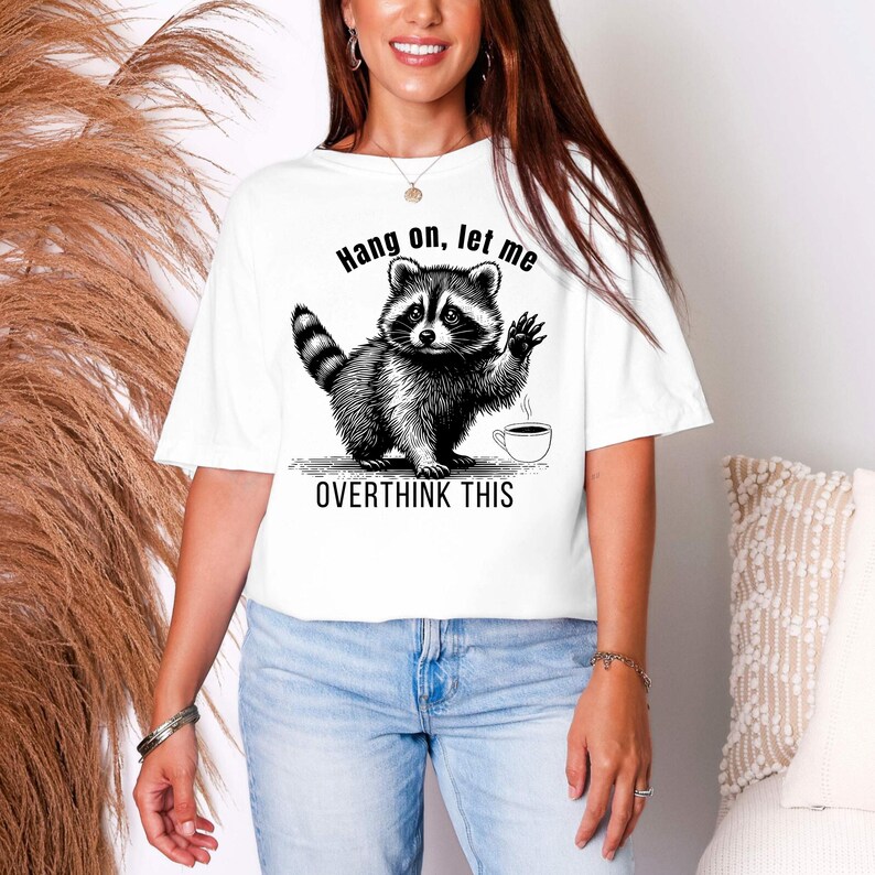 A woman wearing a white t-shirt with a black graphic of a raccoon waving next to a coffee cup and the text "Hang on, let me OVERTHINK THIS."