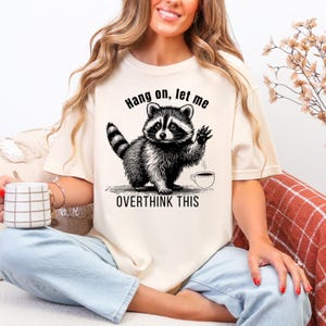 A woman wearing a cream t-shirt with a black graphic of a raccoon waving next to a coffee cup and the text "Hang on, let me OVERTHINK THIS."