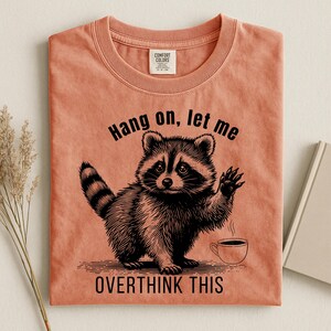 A peach-colored t-shirt with a black graphic of a raccoon holding up a paw next to a steaming coffee cup, with the text "Hang on, let me OVERTHINK THIS."