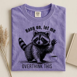A light purple t-shirt with a black graphic of a raccoon holding up a paw next to a steaming coffee cup, with the text "Hang on, let me OVERTHINK THIS."