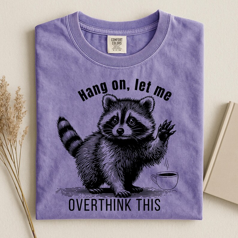 A light purple t-shirt with a black graphic of a raccoon holding up a paw next to a steaming coffee cup, with the text "Hang on, let me OVERTHINK THIS."