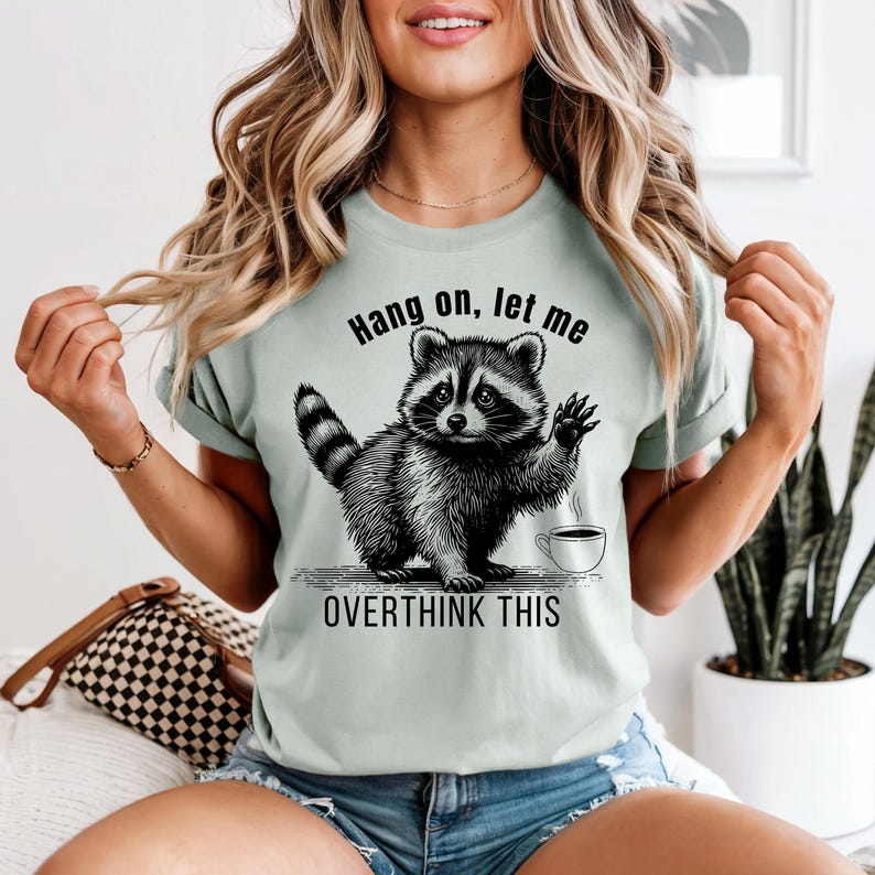 A woman wearing a light green t-shirt with a black graphic of a raccoon waving next to a coffee cup and the text "Hang on, let me OVERTHINK THIS."