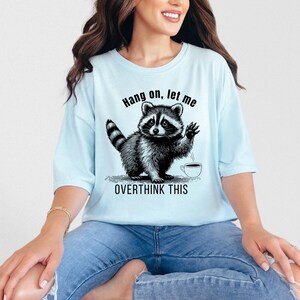 A woman wearing a light blue t-shirt with a black graphic of a raccoon waving next to a coffee cup and the text "Hang on, let me OVERTHINK THIS."