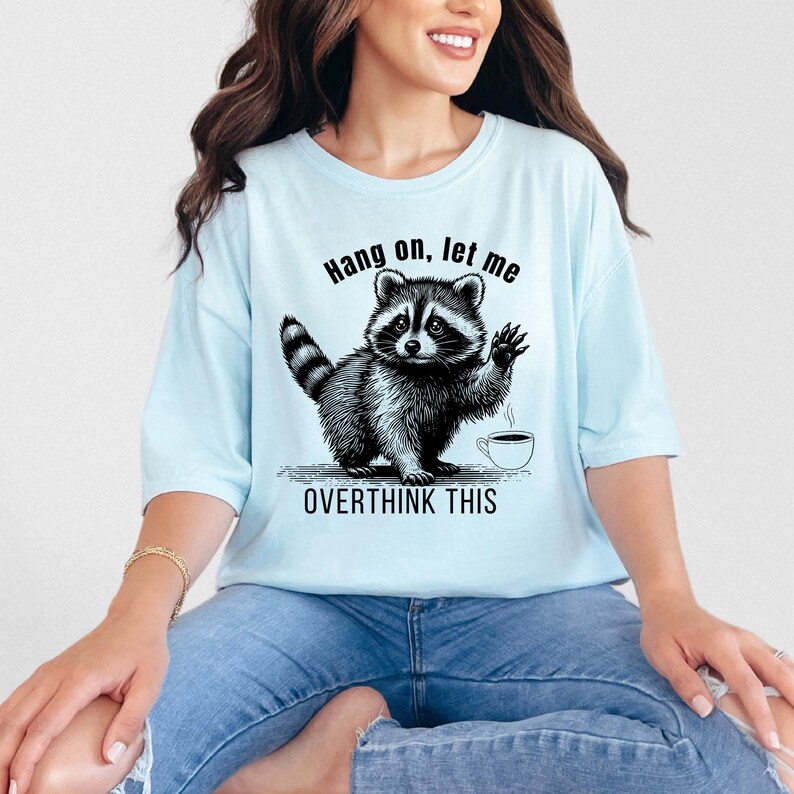 A woman wearing a light blue t-shirt with a black graphic of a raccoon waving next to a coffee cup and the text "Hang on, let me OVERTHINK THIS."