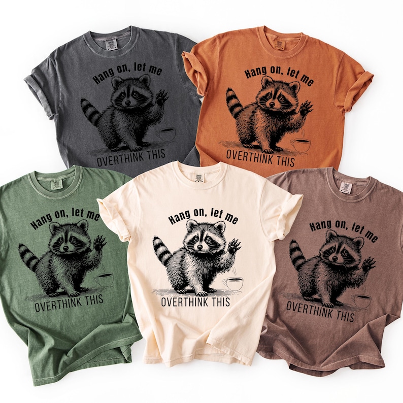Five t-shirts in charcoal, rust, olive, cream, and taupe, each with a black graphic of a raccoon waving next to a coffee cup and the text "Hang on, let me OVERTHINK THIS."