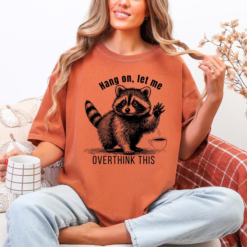 A woman wearing a rust-colored t-shirt with a black graphic of a raccoon waving next to a coffee cup and the text "Hang on, let me OVERTHINK THIS."
